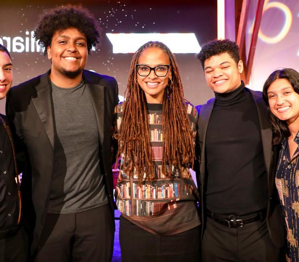 This image features a group of five individuals posing together for a photo. They all appear to be cheerful and are standing in front of a backdrop that includes the word "Juilliard" in large, stylized letters, suggesting that they are at a special event.